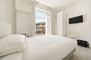 Ein Schlafzimmer mit einem großen Bett, einem an der Wand montierten Fernseher, einem Balkon mit Blick auf Gebäude und einem kleinen weißen Sessel.