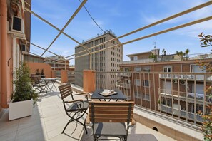 Eine Dachterrasse mit Gartenmöbeln, einem Tisch mit Tasse und Untertasse und Blick auf umliegende Gebäude.