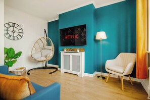 A living room with teal walls, a television, a wicker chair, a white armchair, and a floor lamp. - Blue Serenity Central Oasis | Free Parking | 10PPL (Sheffield)