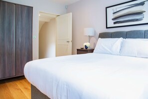 Quarto de hotel com uma cama grande, um guarda-roupa, uma mesa de cabeceira com abajur e um quadro abstrato emoldurado na parede.