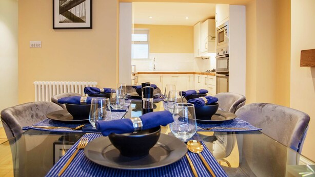 A dining table set with blue napkins, black bowls, and clear glasses, with a kitchen in the background. - Elegant 4-bed Apartment in Pimlico Sleep 7 (London)