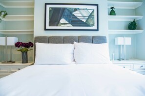 Uma cama bem arrumada com lençóis brancos, uma pintura abstrata emoldurada acima e um vaso com flores na mesa de cabeceira.