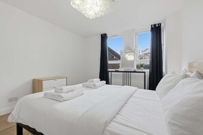 A bedroom with a bed, a bedside table, a mirror, a window with curtains, and a radiator. - Spacious 2-bed Near Tower Bridge Canary Wharf (London)