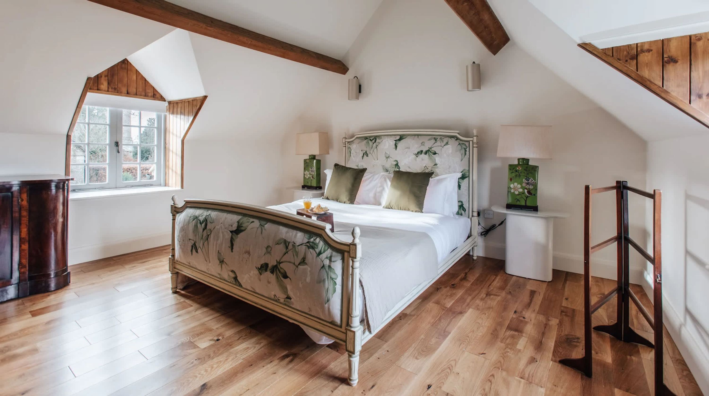 A bedroom with a wooden floor, a bed with floral upholstery, two bedside lamps, and a window with a view.