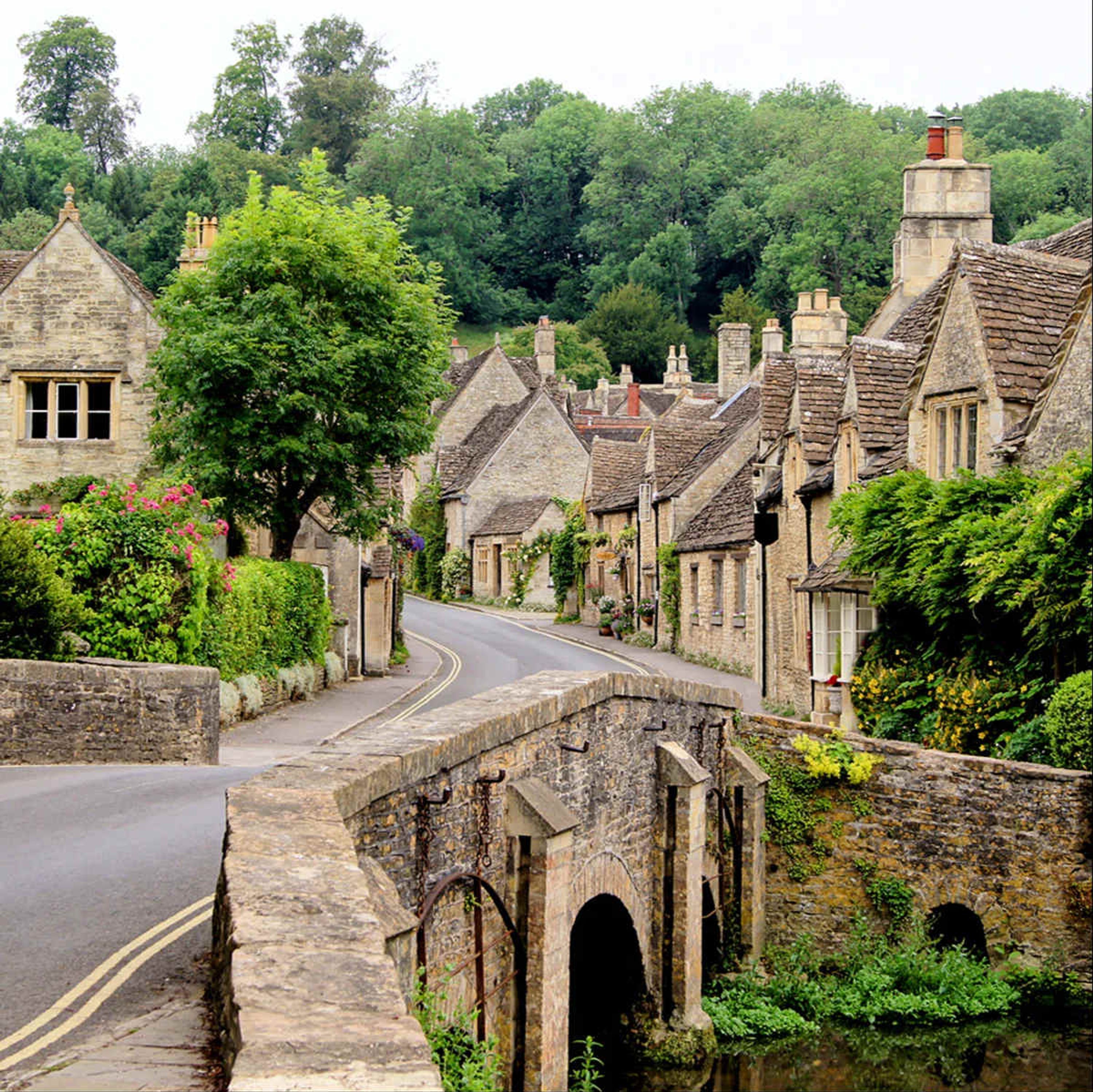 A stone bridge over a narrow road in a village with traditional stone houses, a clear sky, and lush greenery.