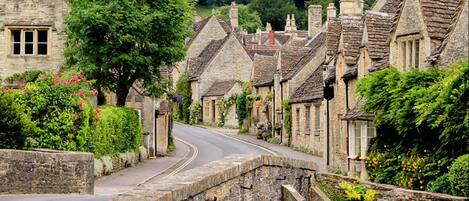 A stone bridge over a narrow road in a village with traditional stone houses, a clear sky, and lush greenery.