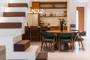 A modern kitchen with wooden cabinets, a dining table with green upholstered chairs, and a staircase with wooden steps. - A Canggu Sojourn Villa (Cemagi)