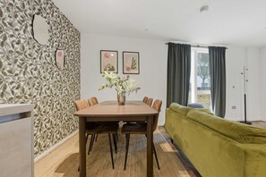 A dining area with a table, chairs, a vase with flowers, a green sofa, and a window with curtains. - Spacious 2BR Near Stratford Station Olympic Park (London)