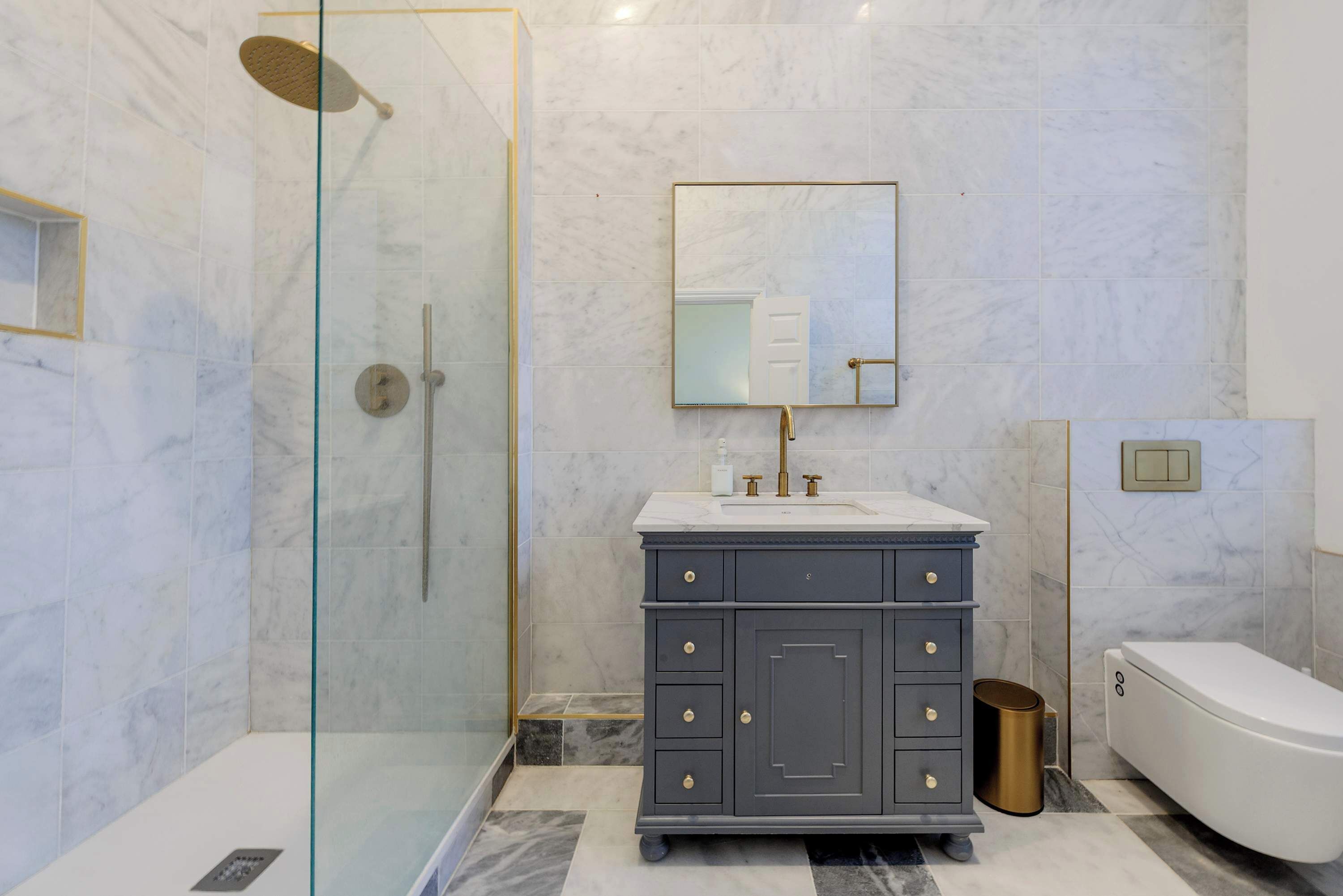 A modern bathroom with a glass-enclosed shower, a marble-tiled wall, a dark vanity with gold handles, and a white toilet.
