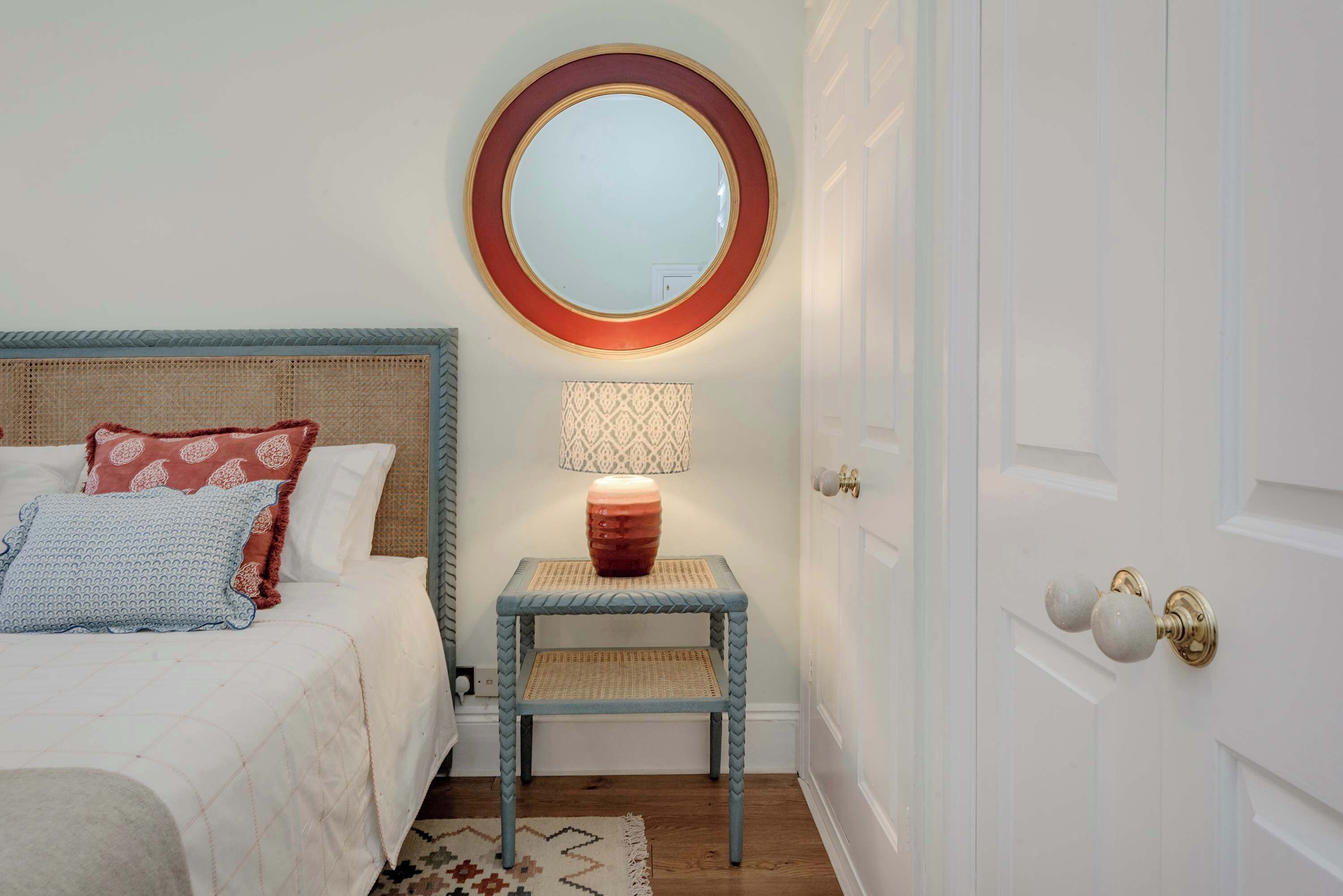 A bedroom with a bed, a bedside table with a lamp, a round mirror, and a door with a knob.