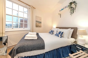 A bedroom with a large window, a bed with a blue skirt, a bedside table with a lamp, and a mirror. - Stylish 2 bed - Tower Hamlets (London)