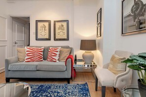 A living room with a grey sofa, red and beige pillows, a blue patterned rug, a white armchair, and framed pictures on the wall. - Stylish 2 bed - Tower Hamlets (London)