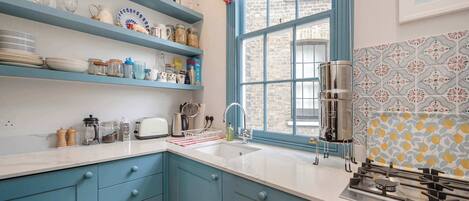Una cocina con armarios azules, encimera blanca, ventana con un valance rojo y azulejos de salpicadero con estampado.