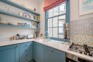 A kitchen with blue cabinets, a white countertop, a window with a red valance, and patterned backsplash tiles. - Gorgeous 3 Bedroom Flat - Pimlico (London)