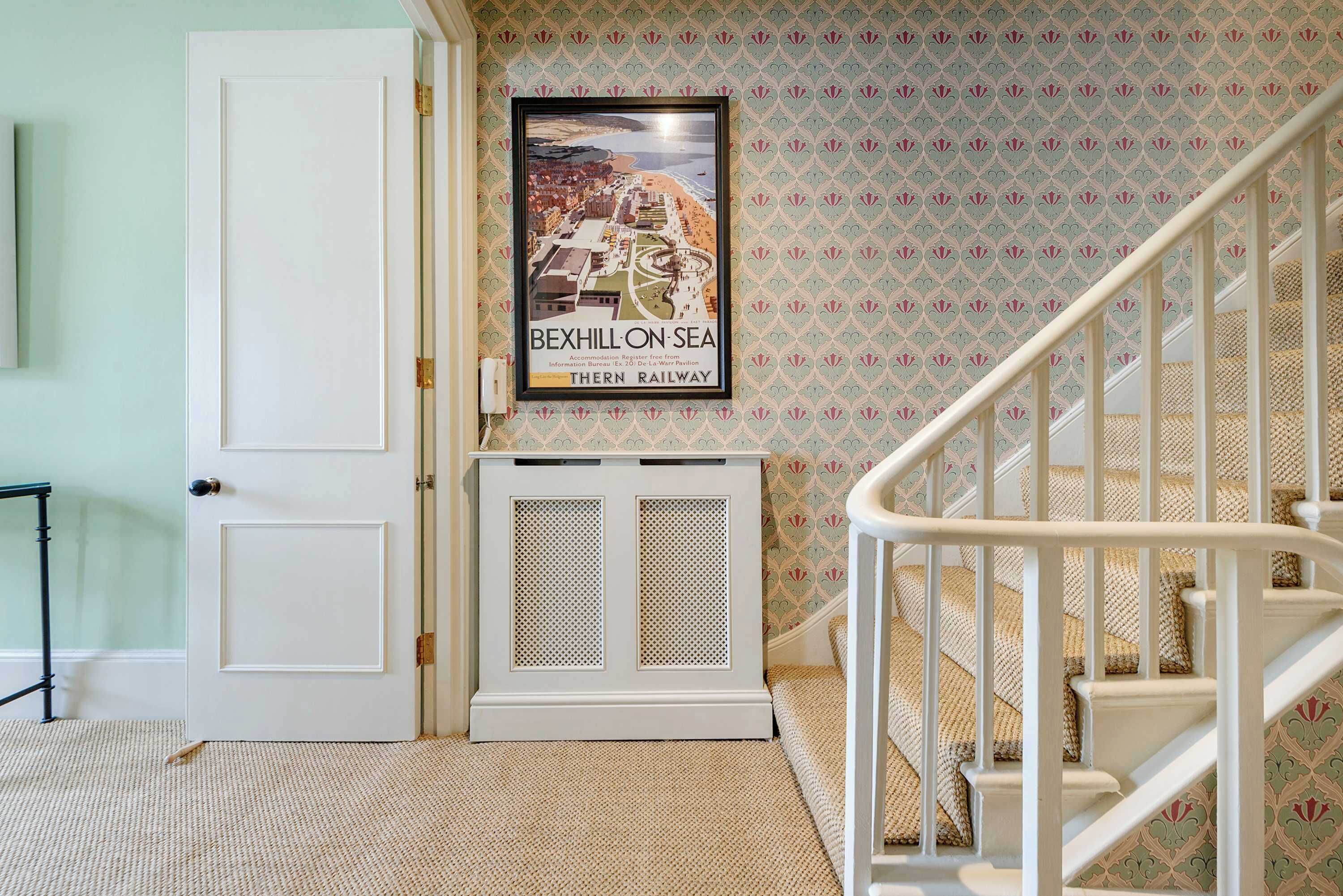 A staircase with a white railing, a framed poster of Bexhill-on-Sea, and a white door with a small table beside it.