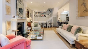 Een ruime woonkamer met een witte bank, een roze fauteuil, een glazen salontafel en een open haard. Een trap leidt naar een eethoek met een witte tafel en stoelen.