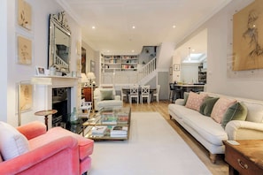 A spacious living room with a white sofa, a pink armchair, a glass coffee table, and a fireplace. A staircase leads to a dining area with a white table and chairs.