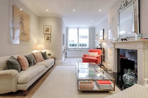 A living room with a sofa, a red chair, a glass coffee table with books, a fireplace, and framed artwork on the walls.