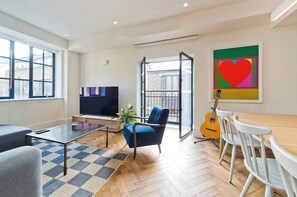 A modern living room with a flat-screen TV, a blue armchair, a wooden dining table with white chairs, a guitar, and a large window. - Elegant 2 Bedroom Flat - King's Cross (London)