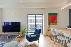 A living room with a TV, a blue armchair, a dining table with chairs, a guitar, and a large window. - Elegant 2 Bedroom Flat - King's Cross (London)