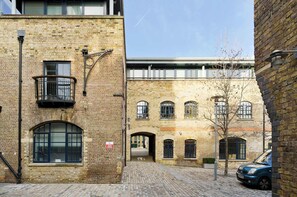 A brick building with arched windows and a balcony, a cobblestone street, and a parked car. - Elegant 2 Bedroom Flat - King's Cross (London)