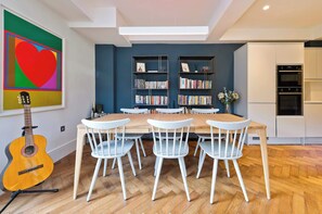 A dining area with a wooden table and white chairs, a bookshelf, and a guitar. - Elegant 2 Bedroom Flat - King's Cross (London)
