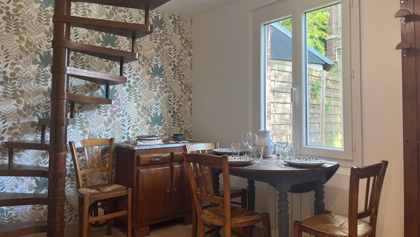 A dining area with a spiral staircase, wooden chairs, a table set for four, and a window with a view of a brick wall and greenery outside.