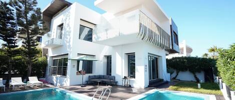 A modern two-story house with a swimming pool, outdoor seating, and a large window overlooking the pool.