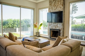 A modern living room with a large sofa, a fireplace, a flat-screen TV, and a view of a swimming pool.