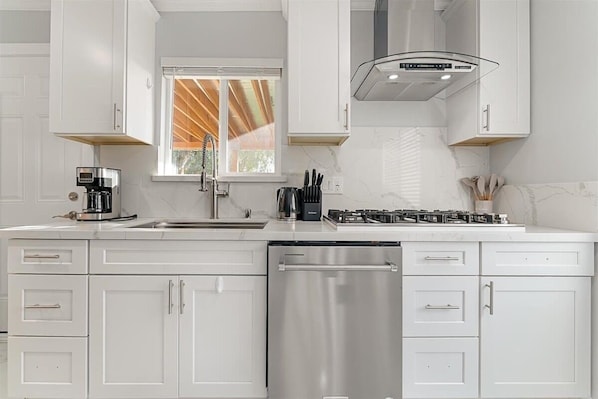 A modern kitchen with white cabinetry, a stainless steel dishwasher, a gas stove, and a window with blinds.