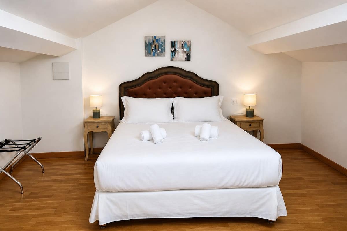 Una cama bien hecha con sábanas blancas, dos lámparas de noche y dos cuadros colgados en la pared.