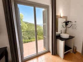 Un baño con lavamanos, espejo y barra para toallas, un cubo de basura y vistas a un balcón con barandilla y vegetación.