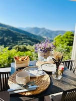 Una mesa dispuesta con platos, vasos y cubiertos, con vistas a montañas y vegetación al fondo.