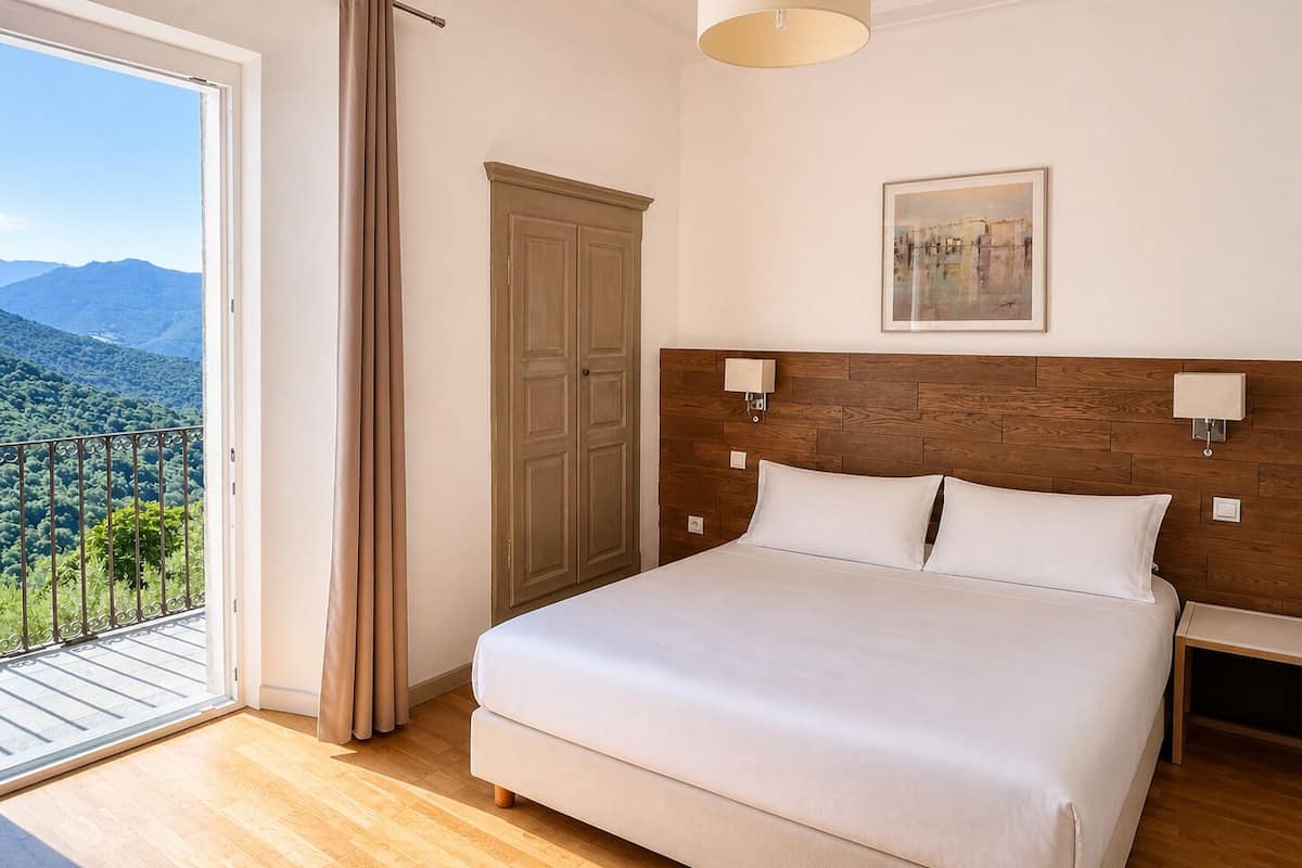 Un dormitorio con cama, mesita de noche, cabecera de madera, ventana con cortinas, puerta y un cuadro en la pared.