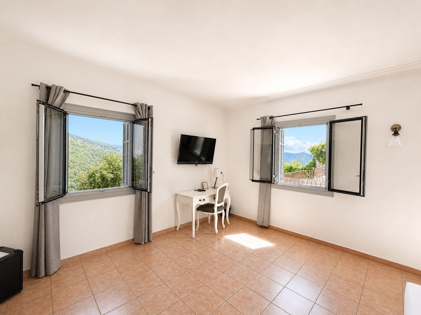 A room with two open windows, a flat-screen TV, a small desk with a chair, and a light fixture.