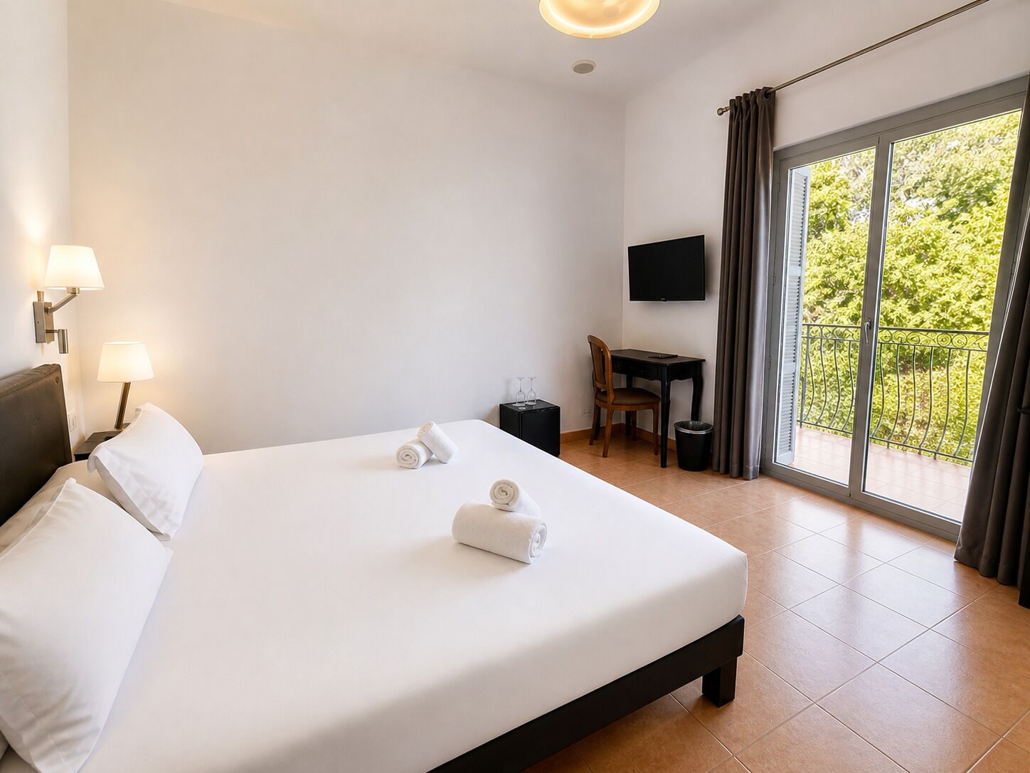 A hotel room with a bed, two bedside lamps, a desk with a chair, a TV, and a balcony with curtains.