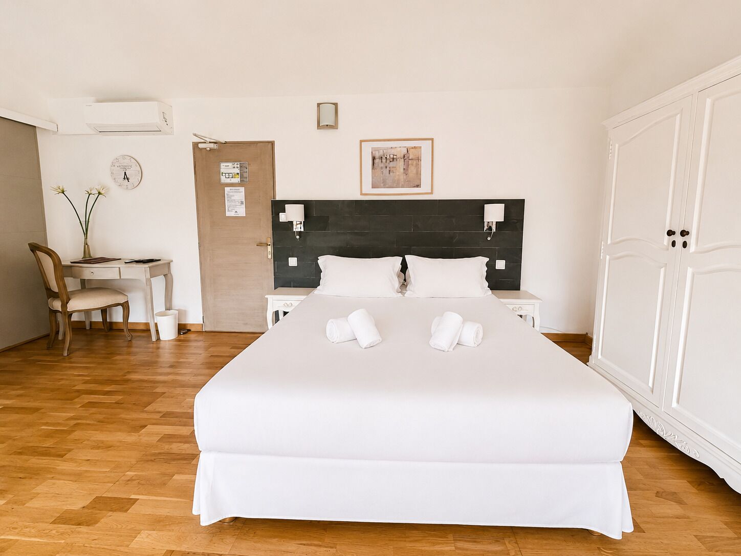 A hotel room with a bed, a desk, a chair, a wardrobe, and a wall-mounted air conditioner.