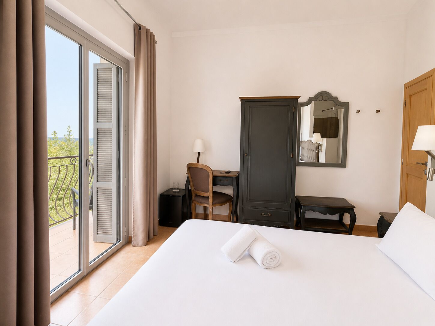 Quarto de hotel com cama, escrivaninha, guarda-roupa e varanda com cortinas.