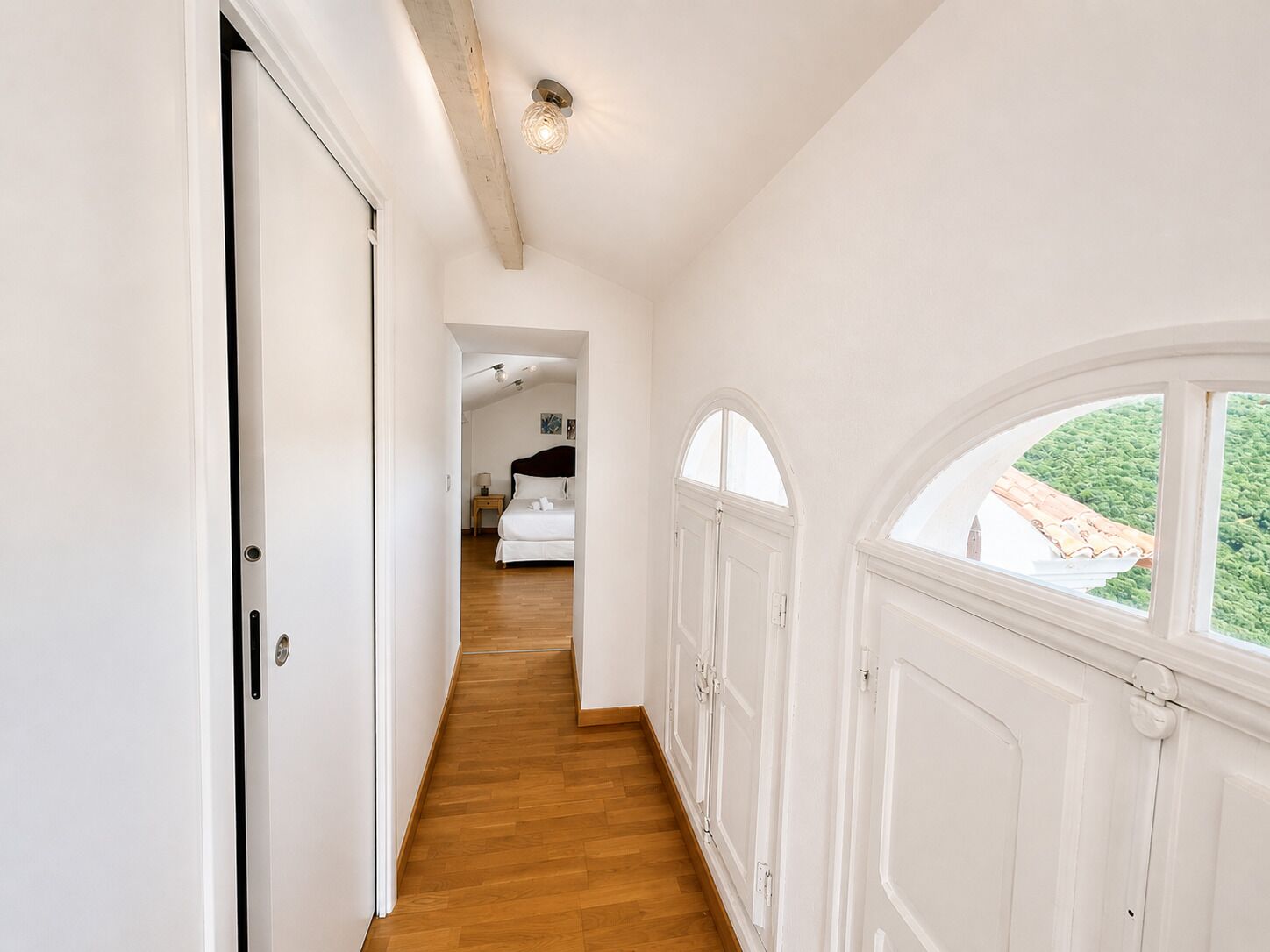 A hallway with a wooden floor, white walls, and a large arched window with a view of greenery.