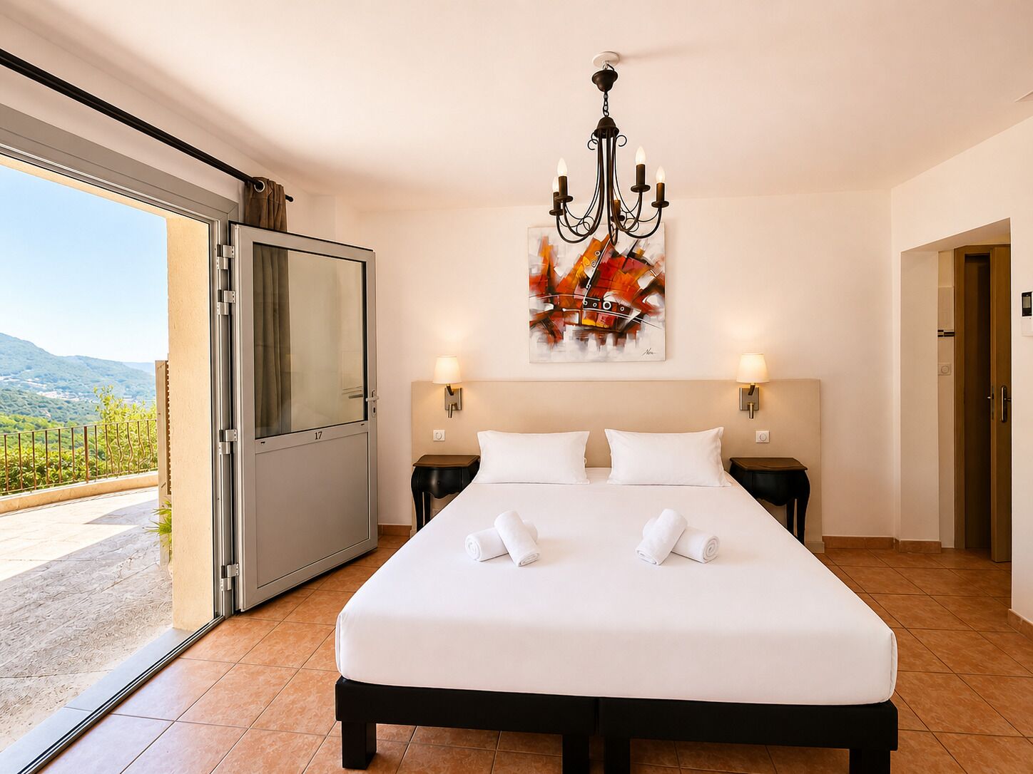 Habitación de hotel con cama, dos lámparas de noche, una araña, un cuadro y una puerta corrediza a un balcón.