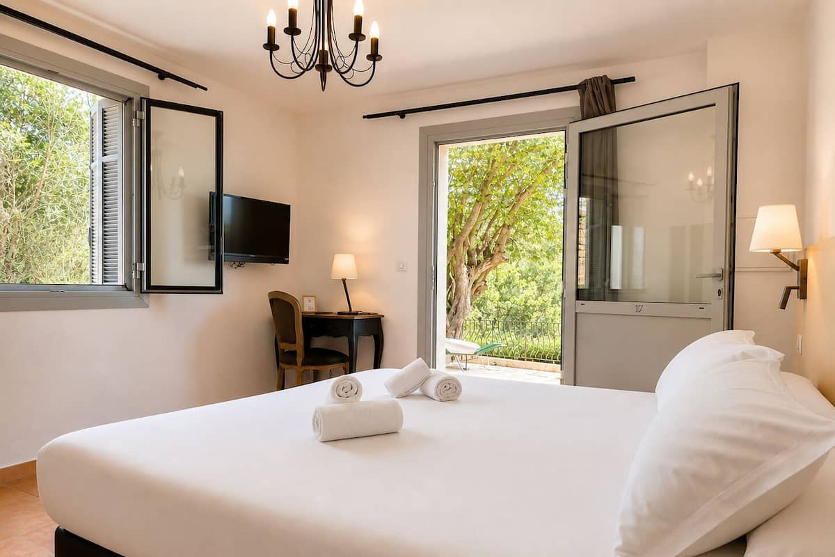 Habitación de hotel con cama, televisión, escritorio con silla, lámpara y vista a zonas verdes.