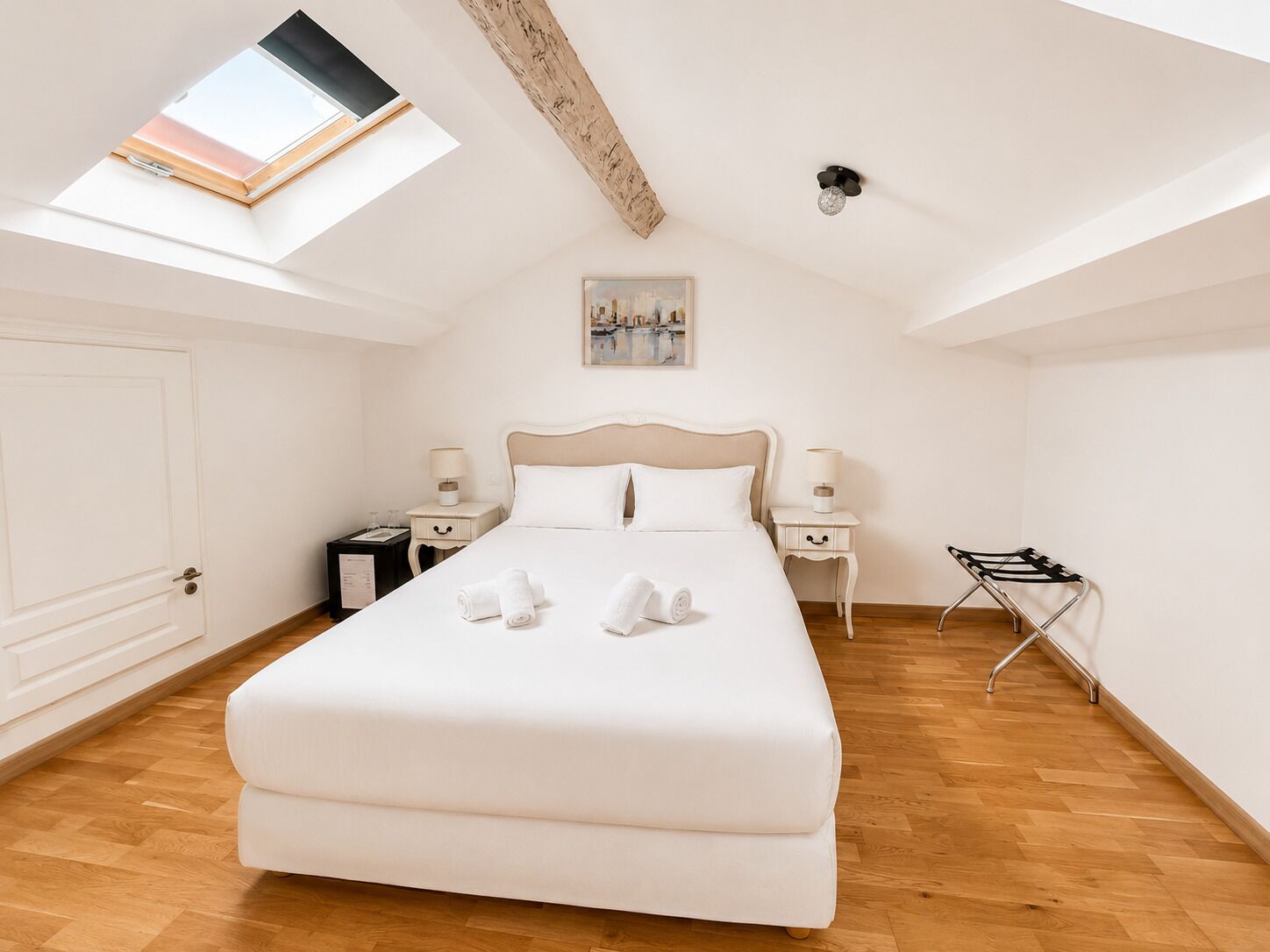 A bedroom with a large bed, two bedside tables, a nightstand, a skylight, a painting, and a folding chair.