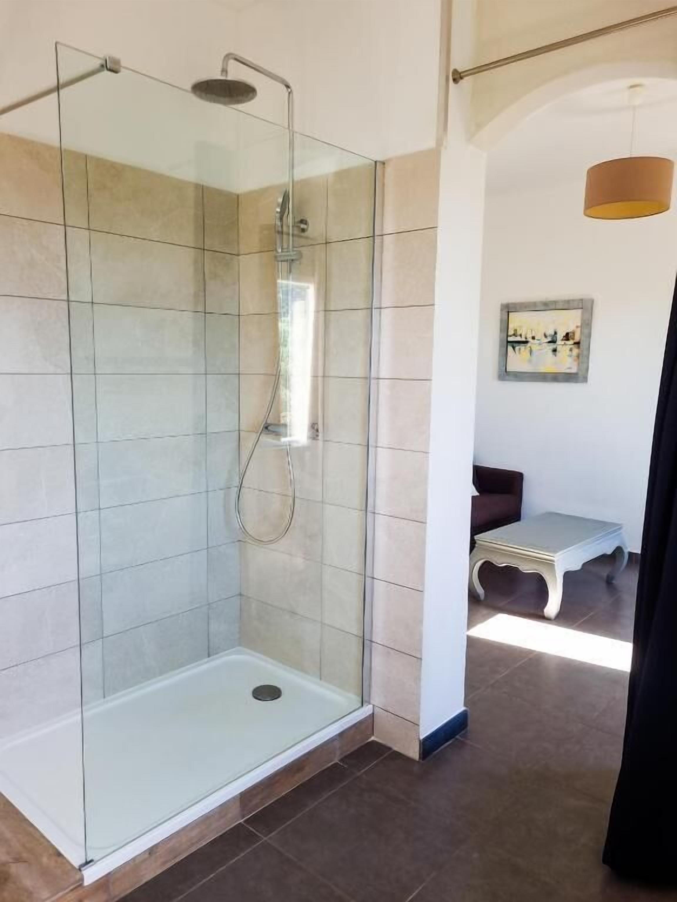 Una cabina doccia in vetro con soffione manuale, un tavolino e una sedia in una stanza con pareti piastrellate e una grande finestra.