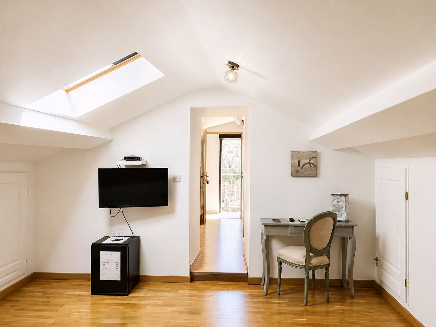 A room with a skylight, a flat-screen TV, a desk with a chair, and a door leading to another room.