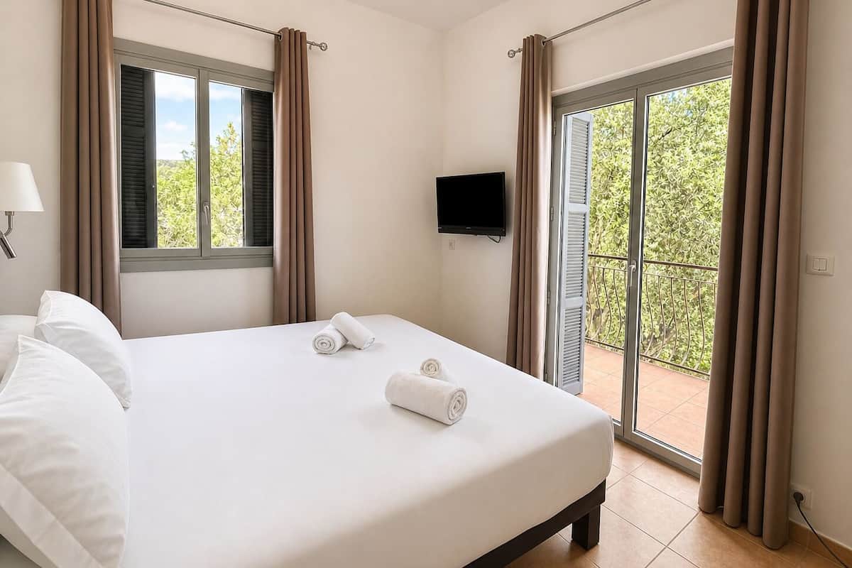 Un dormitorio con cama, dos ventanas con cortinas, televisión y balcón con vistas.