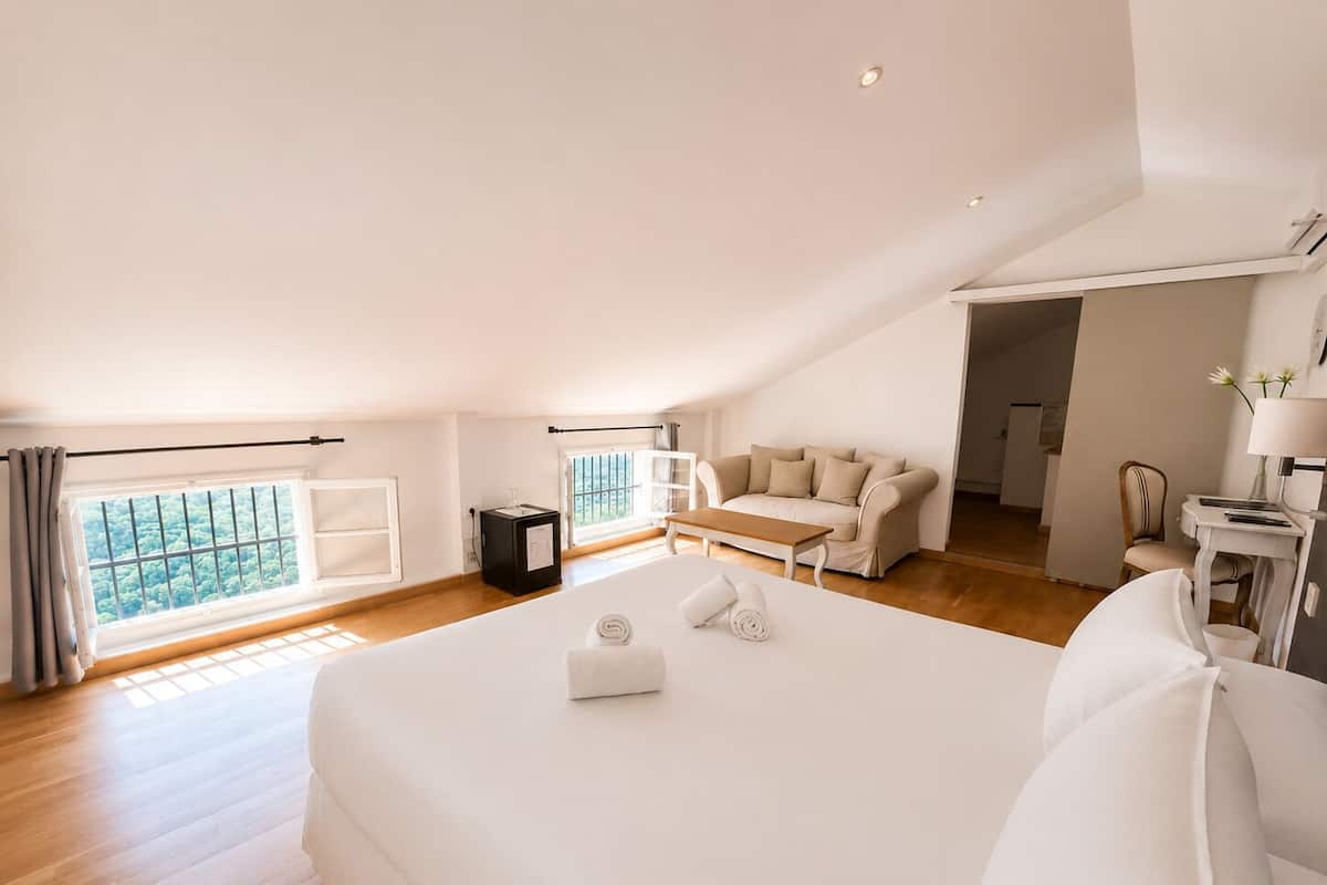 Un dormitorio amplio con una cama grande, un sofá, una mesita y vistas a zonas verdes a través de las ventanas.