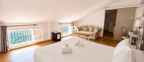 A spacious bedroom with a large bed, a sofa, a small table, and a view of greenery through the windows.