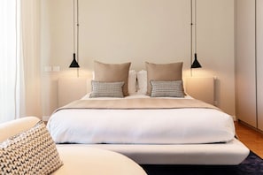 Una cama bien hecha con un edredón blanco y dos almohadas estampadas, flanqueada por dos lámparas colgantes negras.