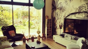 A modern living room with a sofa, armchair, ottoman, and a coffee table. There is a bookshelf and a large window with blinds. - 4.5 km from the beach. Beautiful, quiet house. (Saint-Lormel)