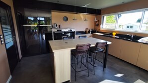 A modern kitchen with a central island, bar stools, a built-in oven, and a large window providing natural light. - 4.5 km from the beach. Beautiful, quiet house. (Saint-Lormel)